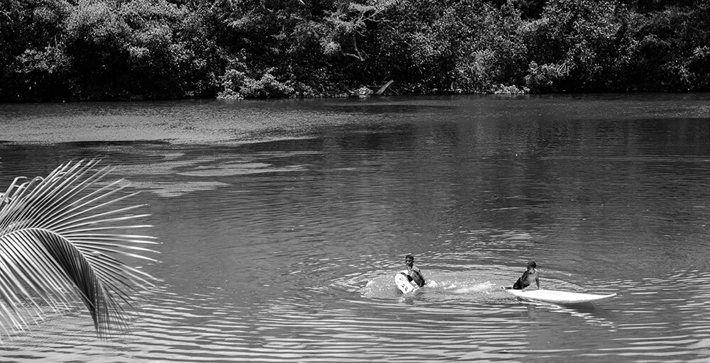 Foto blanco y negro con dos chicos bañándose en un lago con tablas de surf