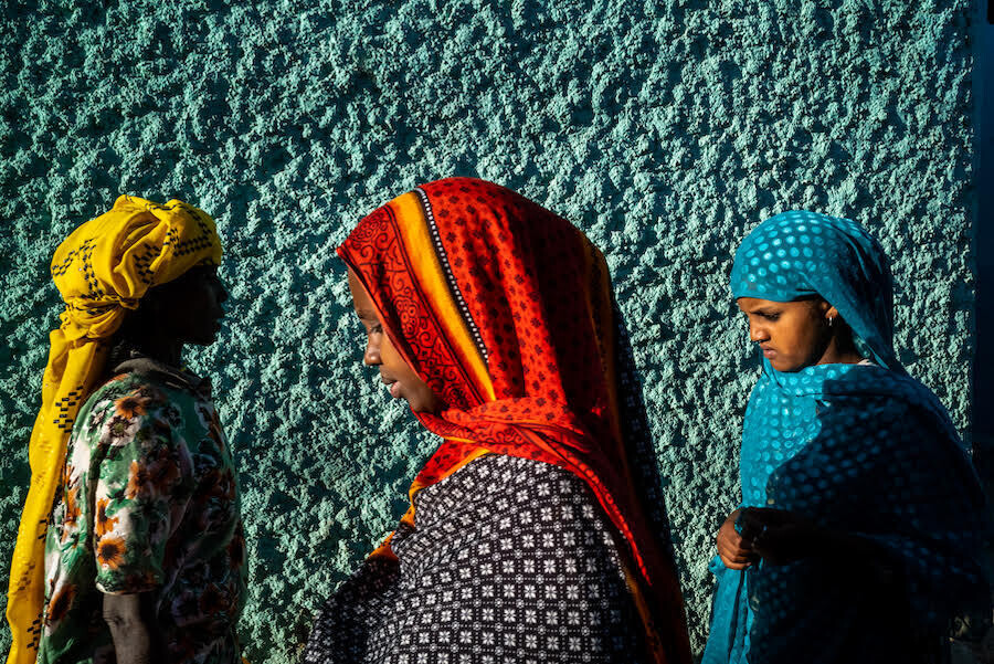 tres mujeres vestidas con pañuelos de vivos colores con una pared color turquesa de fondo