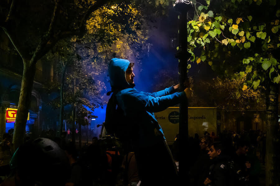 en la escena se ve a un chico iluminado por la luz de una farola a la que está subido y rodeado de gente en penumbra