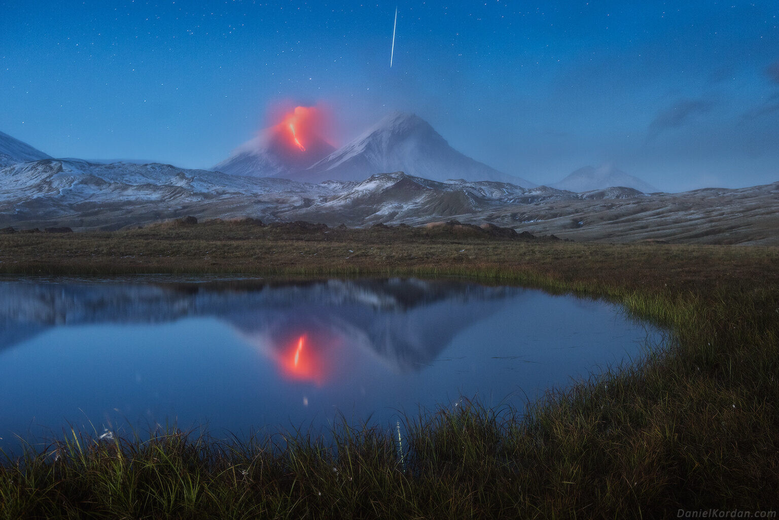 un volcán en erupción y una estrella fugaz en el cielo se reflejan en las aguas de un tranquilo lago en Rusia