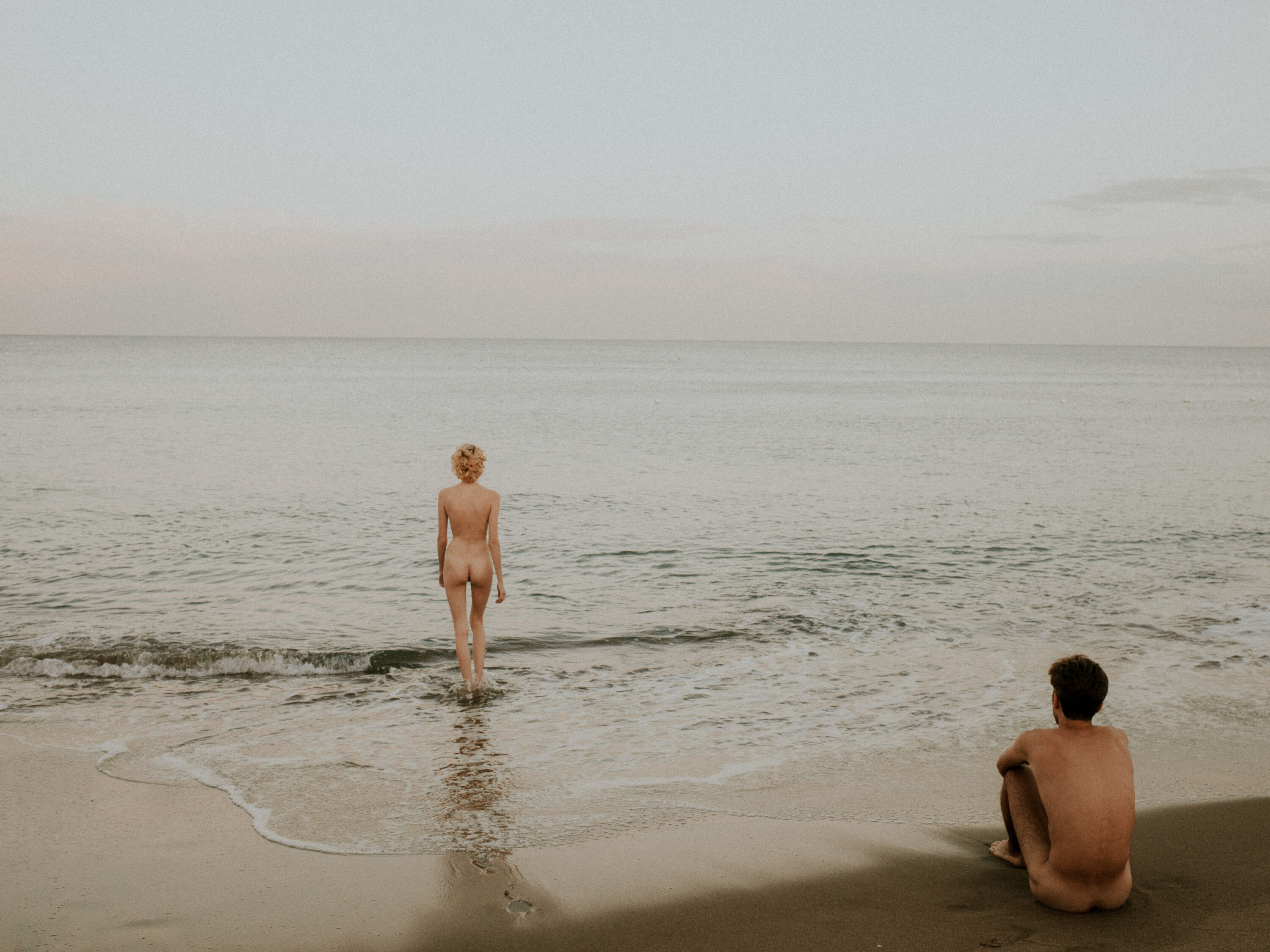 Un chico y una chica en la orilla de la playa desnudos de espaldas.