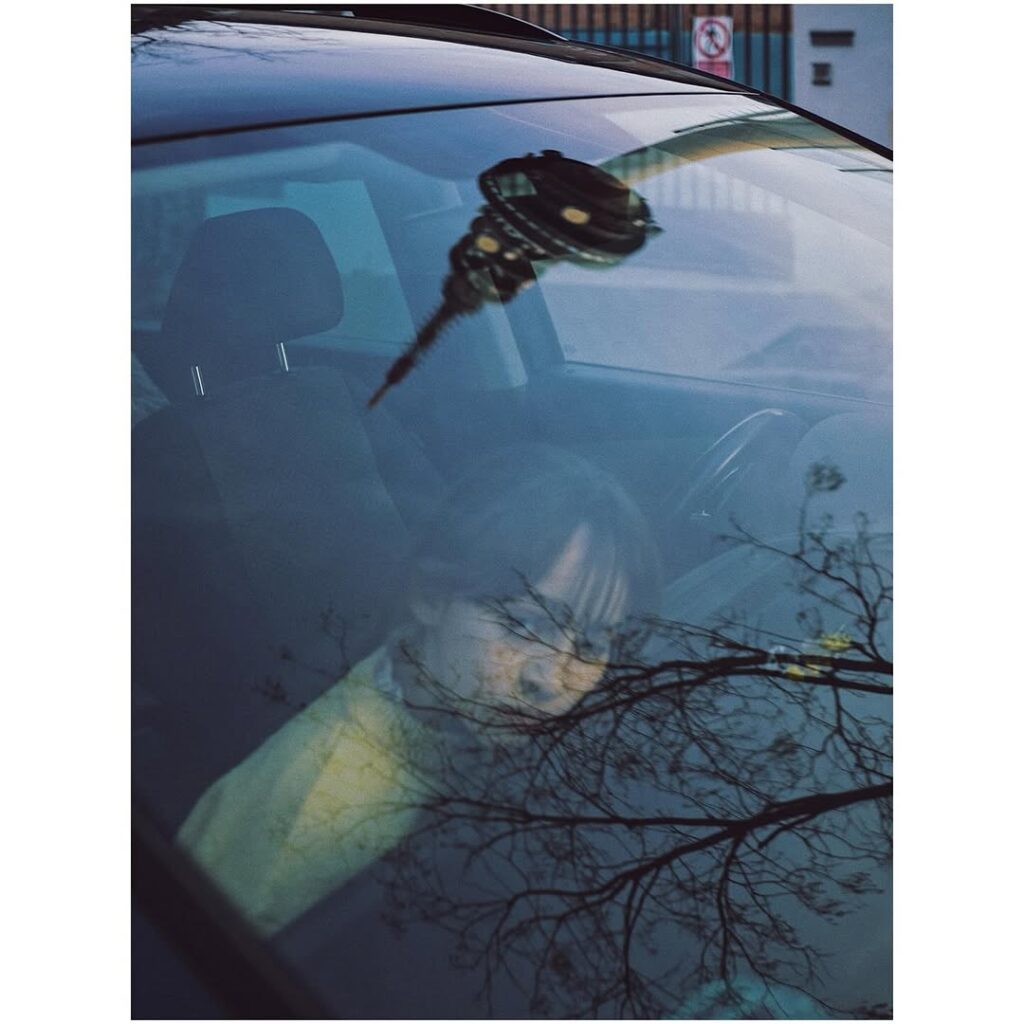 Una niña apoyando la cabeza en el salpicadero de un coche, con el reflejo de un árbol sobre el cristal frontal del coche.